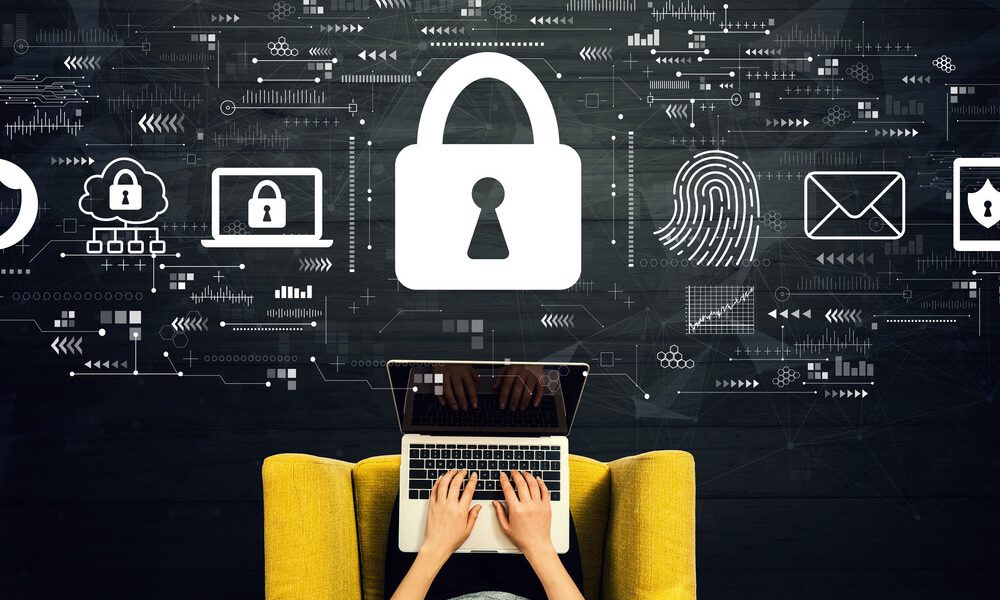 A person typing on a laptop whilst sitting on a yellow chair, with a large padlock and digital security icons floating above, illustrating cyber security.