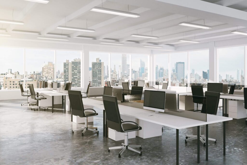 Oficina diáfana con filas de escritorios, sillas ergonómicas y varios monitores de ordenador, con vistas al perfil urbano a través de grandes ventanales.