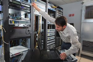 man_datacenter - Informática Lisboa - Informática Lisboa Technician in a data center reaching to rack servers, with a laptop on a rolling cart nearby and network cables in the racks.