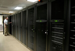 technician_working - Informática Lisboa - Informática Lisboa IT technician in a white shirt stands by a row of tall black server racks in a data center corridor, door open to a cabinet.