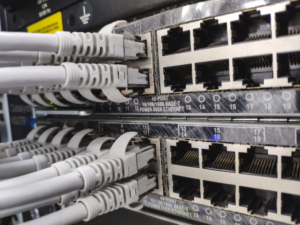 Primer plano de un panel de conexiones de red con varios puertos Ethernet RJ-45 y cables grises conectados a ellos en un armario de servidores, en el que se aprecia el cableado de datos.