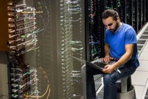 wireless - Informática Lisboa - Informática Lisboa Tech worker in a blue shirt sits beside server racks, using a laptop in a data center to troubleshoot.